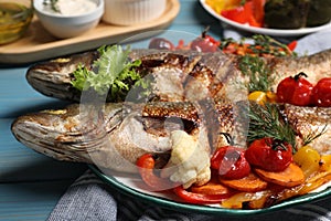 Plate with delicious roasted sea bass fish and vegetables on light blue wooden table, closeup