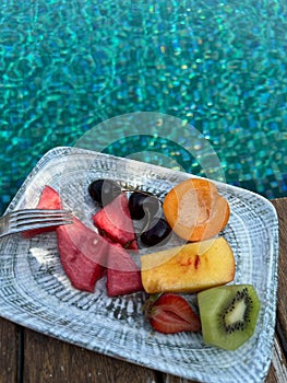 Plate with fresh fruits on wooden deck near outdoor swimming pool. Luxury resort