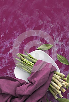 Plate with fresh green asparagus and bay leaves. Purple background and napkin.