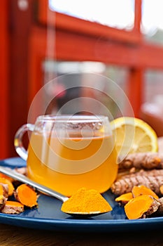Plate with glass cup of hot tea, lemon, turmeric powder and roots on table