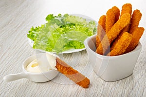 Plate with lettuce, chicken stick in sauce boat with mayonnaise, fried breaded chicken sticks in bowl on wooden table