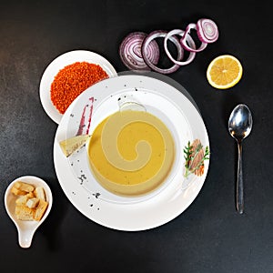 Plate of lentil cream soup with croutons on black table with ingredients for its preparation, Red lentils, cutting onion