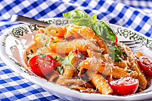 Plate with pasta pene Bolognese sauce cherry tomatoes parsley top and basil leaves on checkered blue tablecloth.