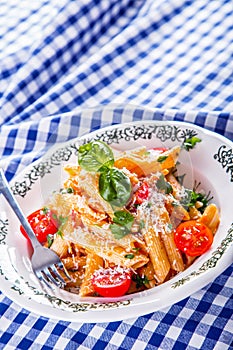 Plate with pasta pene Bolognese sauce cherry tomatoes parsley top and basil leaves on checkered blue tablecloth.