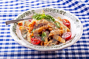 Plate with pasta pene Bolognese sauce cherry tomatoes parsley top and basil leaves on checkered blue tablecloth.