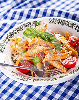 Plate with pasta pene Bolognese sauce cherry tomatoes parsley top and basil leaves on checkered blue tablecloth.