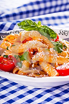 Plate with pasta pene Bolognese sauce cherry tomatoes parsley top and basil leaves on checkered blue tablecloth.