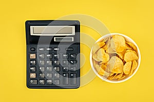 A plate of potato chips and a calculator on a yellow background, top view.