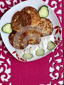 A plate with potato pancakes, sour cream and fresh cucumber, sliced â€‹â€‹with ringlets.