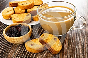 Plate with rusks, bowl with dried grape, transparent cup of coffee with milk on wooden table