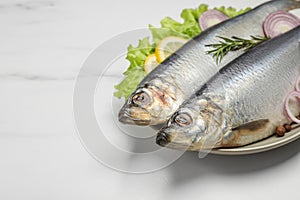 Plate with salted herrings, onion rings, slices of lemon, peppercorns and lettuce on white marble table, closeup. Space for text