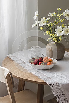 A plate with summer fruits and berries, garden flowers in a ceramic vase on a wooden round table in a cozy living room