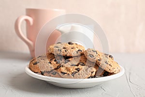 Plate with tasty chocolate chip cookies and blurred cup of milk on gray background
