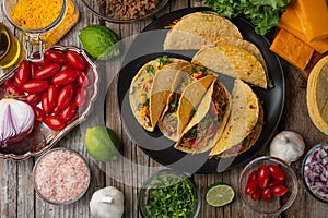 Plate with tasty mexican tacos on rustic wooden table with ingredients for cooking background. Concept of traditional meal. Top