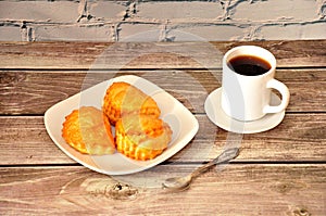 A plate with three cottage cheese buns, a cup of hot coffee on a saucer and a teaspoon on a wooden table