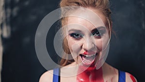 Playful ad predatory young woman grins her teeth with red paint on her neck. Indoors