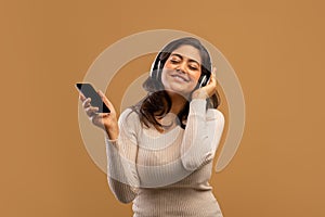 Playful arab lady with headphones and smartphone listening to music and enjoying cool playlist, beige background