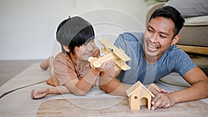 Playful Asian dad playing a wooden figure model with his son while laying on the living room floor