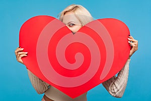 Playful blond girl peeping from big paper red heart in her hands, saint valentines day celebration, love and fondness