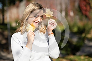 Playful blond woman white sweater, holding golden foliage yellow leaves maples in two hands,hiding face, blur. Close up