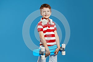 Playful cool kid with headphones wearing trendy outfit, holding skateboard against blue background