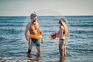 Playful couple in water at beach. Man with rubber duck float and diving googles, looking at woman with water gun. Holiday,