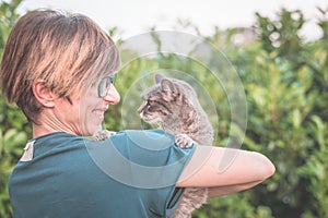 Playful domestic cat held and cuddled by smiling woman with eyeglasses. Outdoor setting in green home garden. Shallow depth of fie