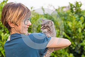 Playful domestic cat held and cuddled by smiling woman with eyeglasses. Outdoor setting in green home garden. Shallow depth of fie