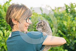 Juguetón doméstico gato sostuvo a abrazo de acuerdo a mujer sonriente anteojos. externo configuración en verde jardín. poco profundo profundidad de puaj 