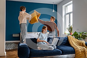 Playful european siblings boy and girl pillow fighting distracting busy mom relaxing on sofa at home