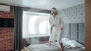 Playful guy in bathrobe dancing on bed listening to music through headphones in hotel