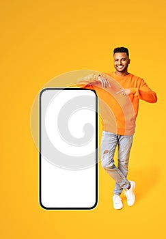 Playful handsome indian man pointing finger at huge smartphone with empty blank screen isolated on yellow