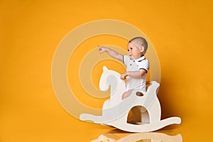 Playful infant baby boy toddler is riding his wooden horse for children and pointing his finger at free copy space