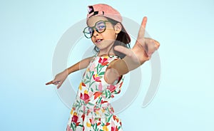 Playful kid in summer dress and pink cap pointing at the camera by fingers, having fun. The little girl directing fingers at the