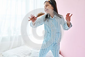 Playful little girl in pajama on the bed holding a hair brush like microphone singing imitates herself a real singer in the