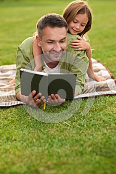 Playful little girl watching her dad reading a book while lying on a blanket. Happy young father and his daughter having