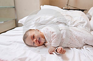Playful lovely baby boy in white clothes, smiling and laughing lying on white bedsheets on the bed at home