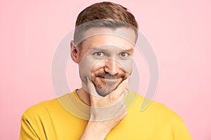 Playful man with sly insidious look touching chin with hand looking at camera on pink background.