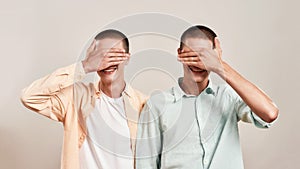 Playful mood. Two cheerful twin brothers covering eyes with hands and smiling while posing together isolated over beige