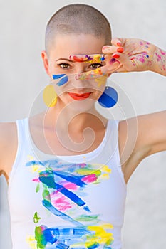 Playful portrait of a young gorgeous female artist painter covered in paint, looking and smiling at camera through her fingers.
