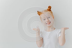 Playful red haired girl raises palms, has double buns, looks happily at camera, wears casual white t shirt, poses indoor, makes