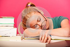 Playful schoolgirl with two hair tails and big eyeglasses sitting behind the small table full of books fall asleep while studying.