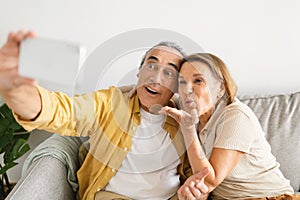 Playful senior spouses making selfie on phone, woman blowing air kiss to webcam, capturing moment with husband at home