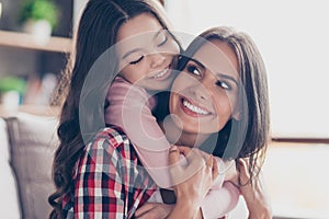 Playful small girl with long dark hair is hugging her mum`s neck