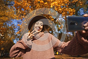 Playful smiling woman in treny hat posing for camera phone at fall season park