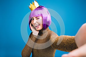 Playful stylish selfie portrait brightful young woman with cut purple hair, golden crown showing tongue on blue