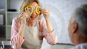 Playful woman making faces with pepper slices, senior couple having fun at home
