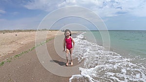 Playful young girl, kid running barefoot in pink swimwear on sandy beach in summer vacation, sea waves approaching the shore