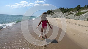 Playful young girl, kid running barefoot in pink swimwear on sandy beach in summer vacation, sea waves approaching the shore