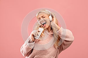 Playful young lady singing and holding smartphone like microphone wearing wireless headphones, pink background